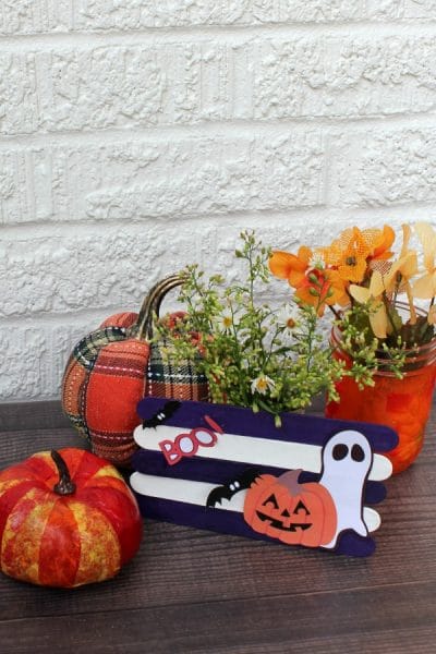 Halloween decoration with purple and white popsicle stick fence. White ghost, black bats, orange pumpkin images glues on popsicle sticks.