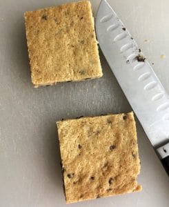 Two squares of chocolate chip cookie dough with a sharp knife on a white cutting board.