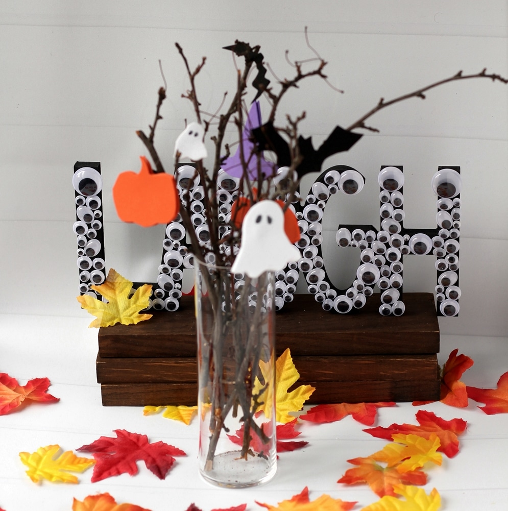 Halloween Tree made from branches, halloween ornaments made from paper in a clear glass vase surrounded by leaves.
