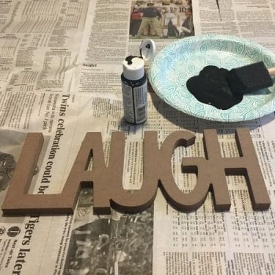 Laugh letters in a wood block. Blue paper plate with foam paint brush and bottle of black paint.